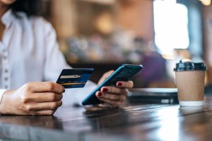 Person making an online purchase using a smartphone and a debit card at a coffee shop, highlighting the importance of secure financial transactions.