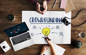 Top view of a workspace with a laptop, notebook, and a paper showing a lightbulb surrounded by dollar signs, illustrating the concept of crowdlending and collaborative funding strategies.