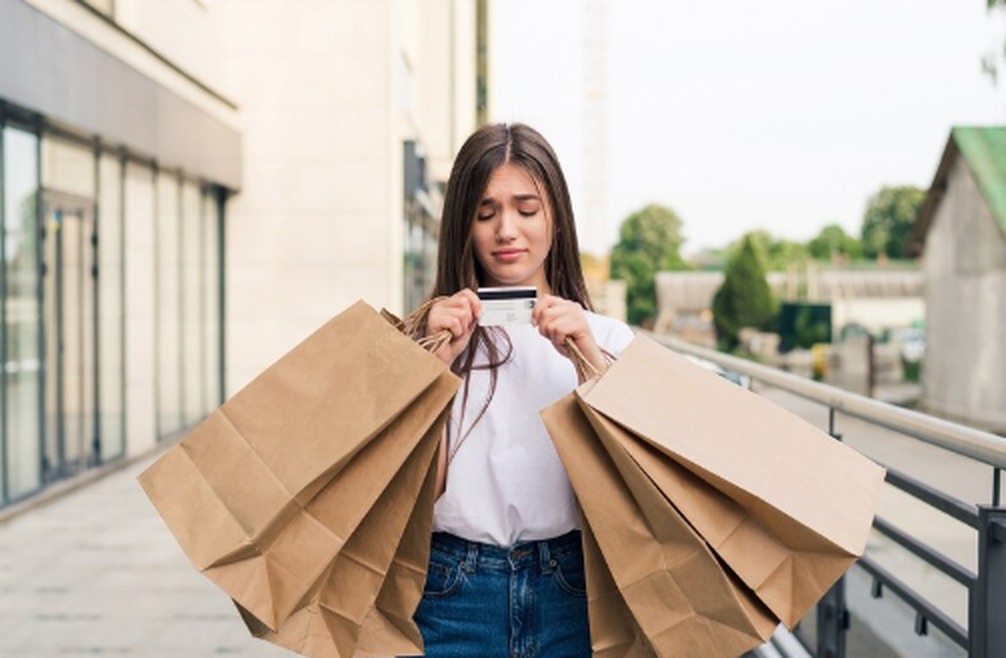 Young woman overwhelmed by impulse buying, holding multiple shopping bags and looking worriedly at her credit card after making unplanned purchases.