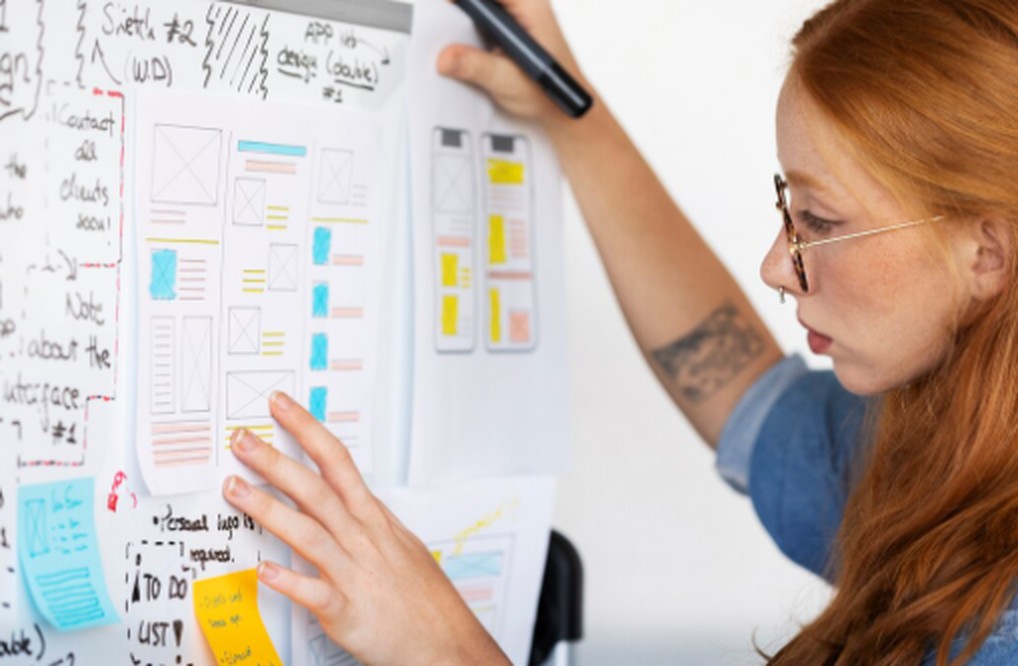 A focused woman reviews a design layout on a whiteboard, analyzing app interface sketches and color-coded notes to improve workflow and optimize saving strategies in a creative project.