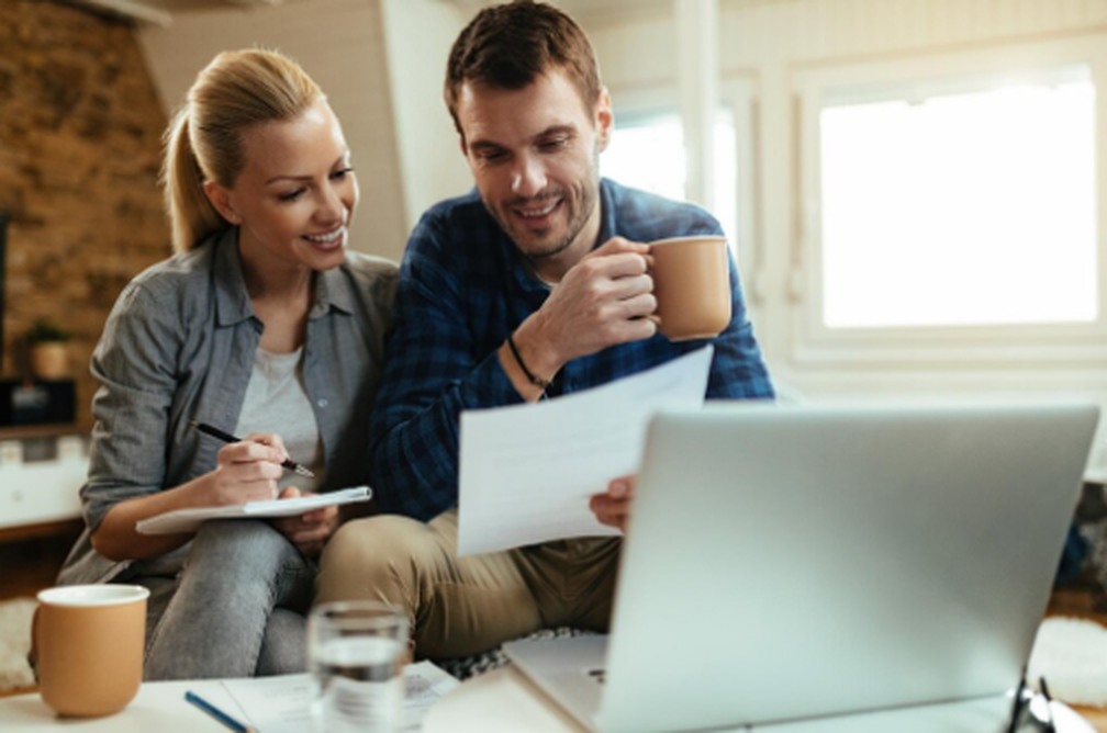 A smiling couple sitting together at a table, reviewing documents and taking notes on a laptop, illustrating a financial checklist for couples planning their budget and expenses.