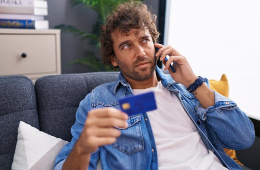 Man sitting on a couch holding a blue card while talking on the phone, appearing thoughtful about renegotiating credit card debt.