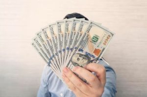 Person holding a fan of hundred-dollar bills in front of their face, symbolizing the growing trend of cash payments as an alternative to credit cards.