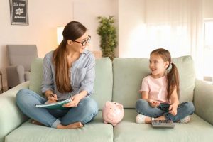 A mother teaching her daughter about money at home, with a piggy bank and calculator on the sofa, illustrating how multigenerational families manage financial education together.