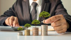 A person in a suit stacking coins with small trees growing on top, symbolizing how microinvestments can grow wealth and promote long-term financial growth.