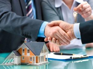 Business handshake in front of a small house model and contract documents, symbolizing an agreement or transaction in the real estate market.